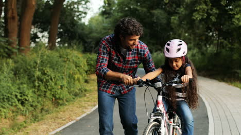 Portrait-of-a-little-girl-and-her-father.-Dad-teaching-his-daughter-to-ride-a-bike.-Smiling.-Moving-camera.-Blurred-background